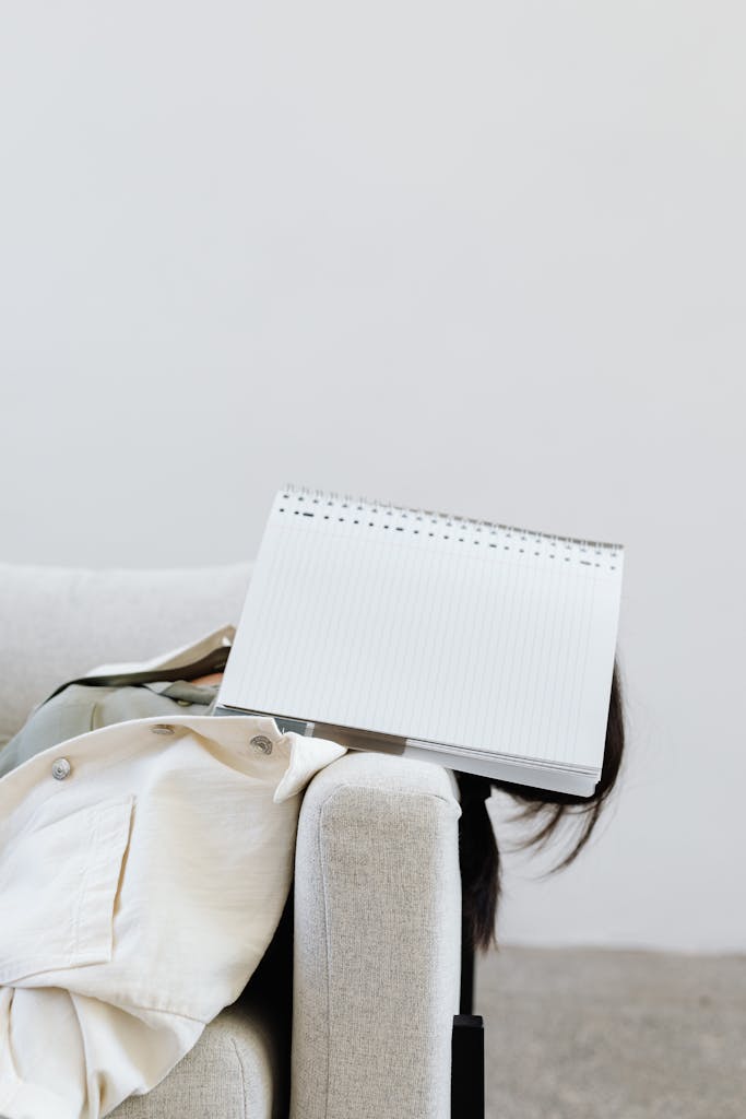 A woman rests with a notebook on her face in a modern office setting, showing signs of tiredness.
